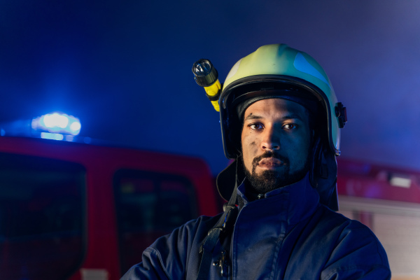 A portrait of dirty firefighter man on duty with fire truck in background at night, smiling.