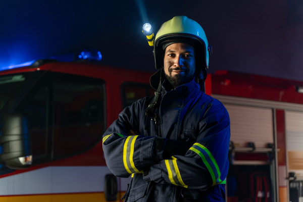 A portrait of dirty firefighter man on duty with fire truck in background at night, smiling.