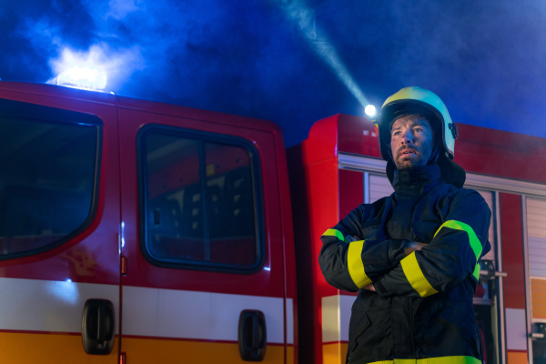 A low angle view of m firefighter talking to walkie talkie with fire truck in background at night.