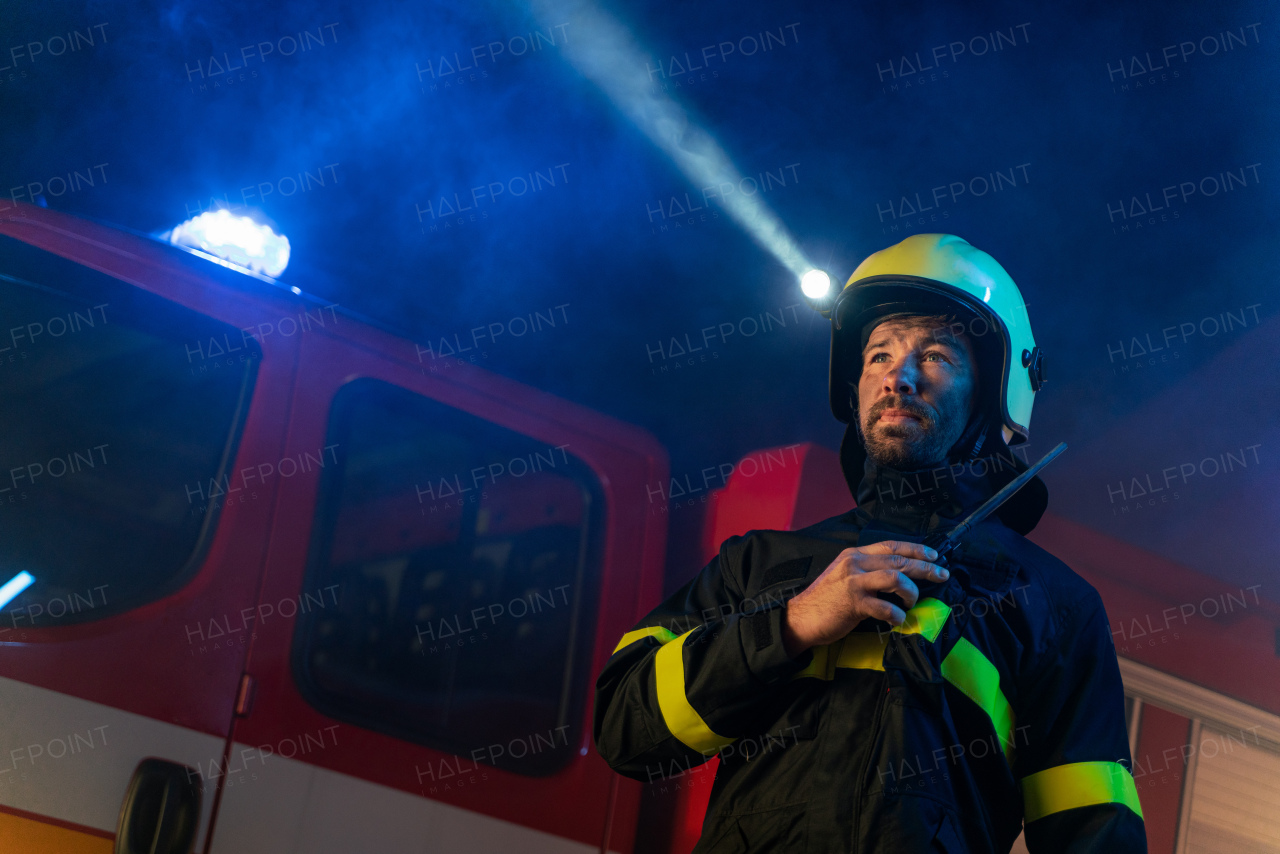 A low angle view of m firefighter talking to walkie talkie with fire truck in background at night.