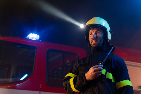 A low angle view of m firefighter talking to walkie talkie with fire truck in background at night.