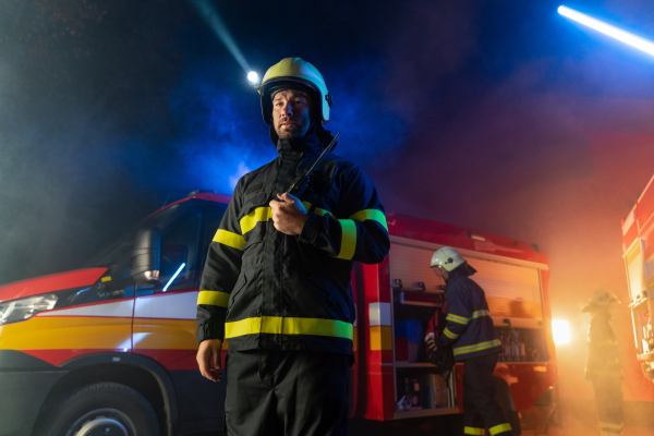 A low angle view of m firefighter talking to walkie talkie with fire truck in background at night.