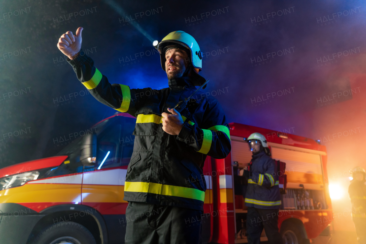 A low angle view of m firefighter talking to walkie talkie with fire truck in background at night.