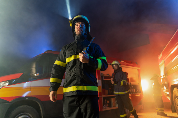 A low angle view of m firefighter talking to walkie talkie with fire truck in background at night.