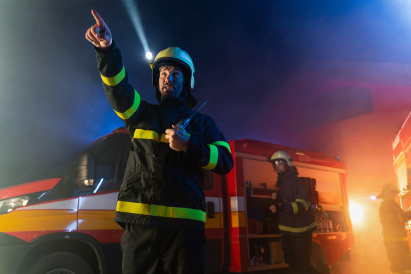 A firefighter talking to walkie talkie with fire truck in background at night.