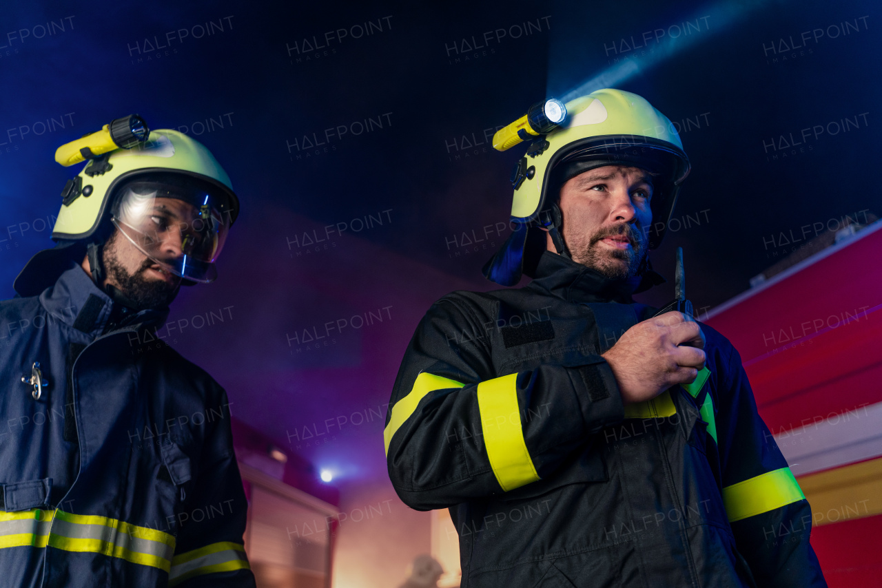 A firefighter talking to walkie talkie with fire truck in background at night.