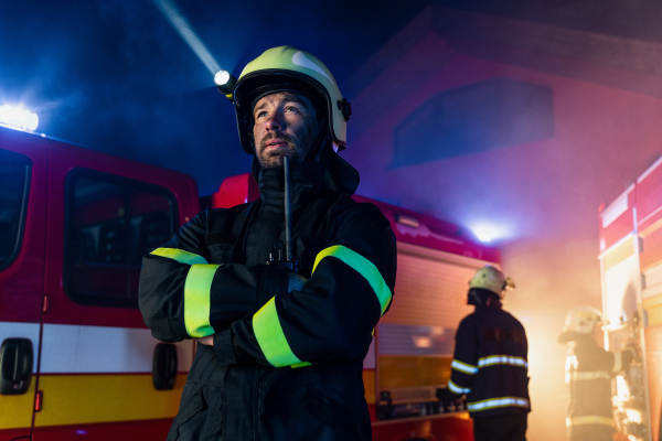 A low angle view of m firefighter talking to walkie talkie with fire truck in background at night.