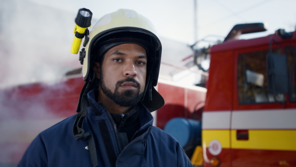 A young American-African firefighter man looking at camera with fire truck and smoke in background