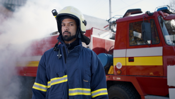 A happy young American-African firefighter man looking at camera with fire truck and smoke in background