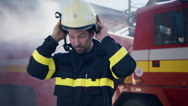 A mature dirty firefighter putting on helmet with fire truck at background