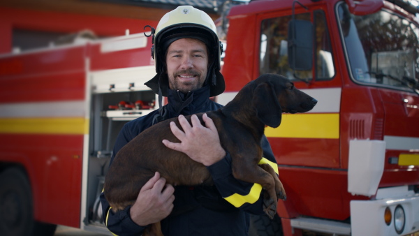 A happy mature firefighter man holding dog and looking at camera with fire truck in background