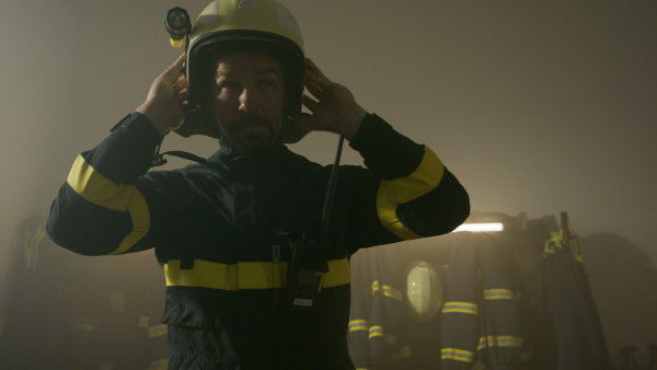 A mature caucasian firefighter putting on helmet indoors in fire station, getting ready for action at night