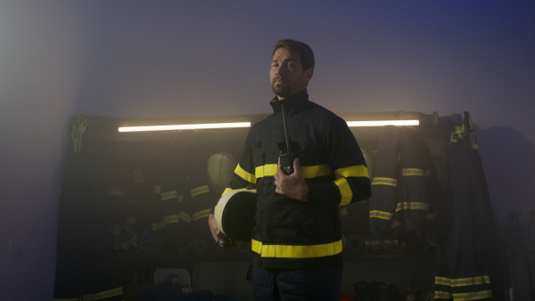 A mature firefighter man hodling walkie talkie and looking at camera indoors in fire station at night.