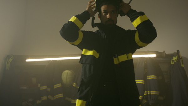 A young African-American firefighter putting on helmet and talking to walkie talkie in fire station.