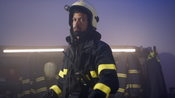 A young African-American firefighter putting on helmet and looking at camera in fire station at night.