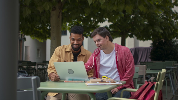 A young man with Down syndrome with his mentoring friend sitting outdoors in cafe and studying.