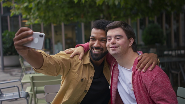 A young man with Down syndrome and his mentoring friend sitting and taking selfie outdoors in cafe