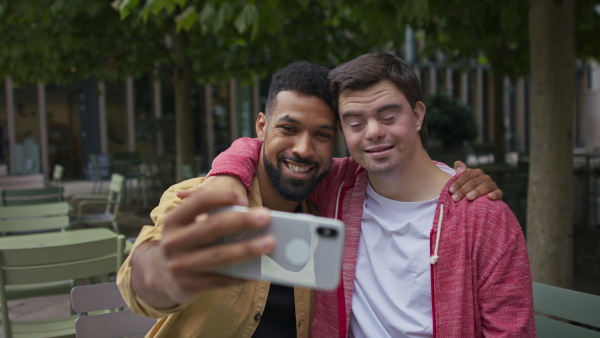 A young man with Down syndrome and his mentoring friend sitting and taking selfie outdoors in cafe, making faces