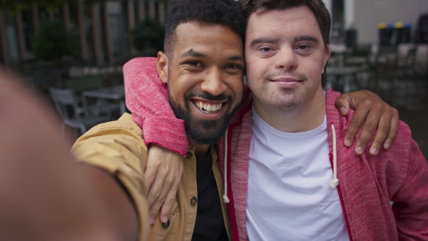 A young man with Down syndrome and his mentoring friend looking at camera and taking selfie outdoors in cafe