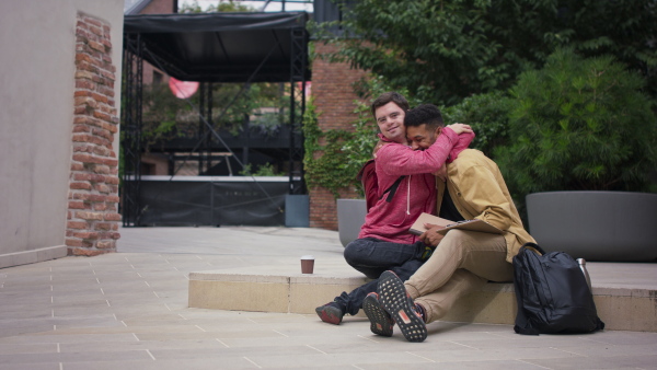 A happy young man with Down syndrome and mentoring friend sitting and hugging outdoors