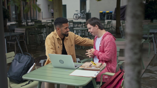 A young man with Down syndrome with his mentoring friend sitting outdoors in cafe and studying.