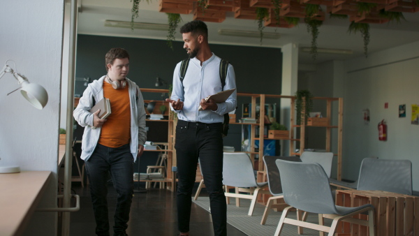 A young man with Down syndrome and his tutor walking together indoors at school.