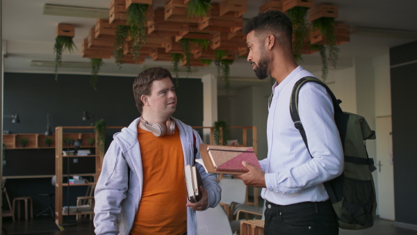 A young man with Down syndrome and his tutor standing and talking together indoors at school.