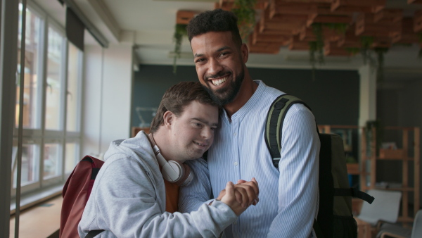 A young man with Down syndrome and his tutor greeting and looking at camera indoors at school.