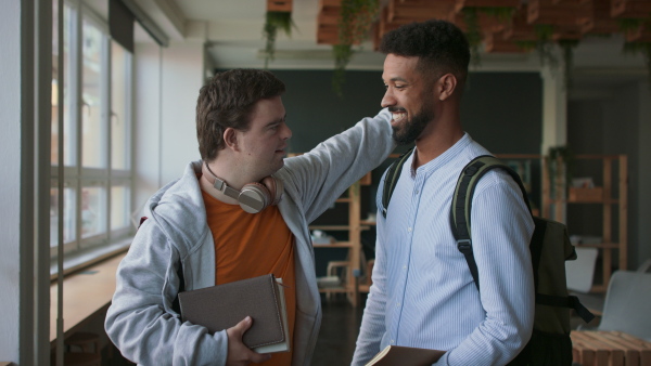 A young man with Down syndrome and his tutor hugging and looking at camera indoors at school.