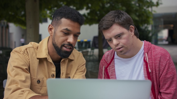 A young man with Down syndrome with his mentoring friend sitting outdoors in cafe and using laptop.