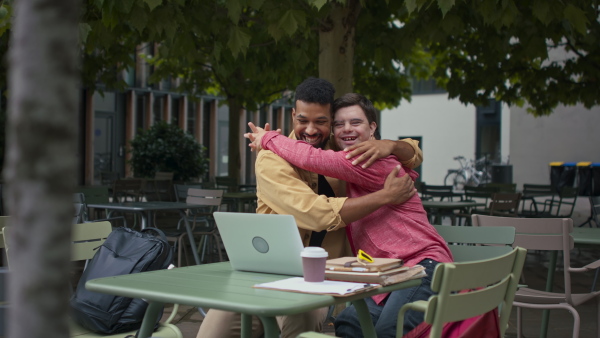 A young man with Down syndrome with his mentoring friend sitting outdoors in cafe, hugging.