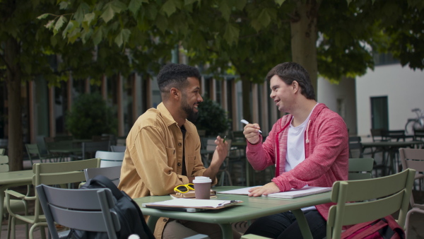 A young man with Down syndrome with his mentoring friend using smartphone and sitting outdoors in cafe.