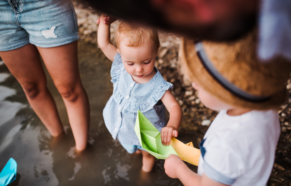 A midsection of young family with two toddler children outdoors by the river in summer, playing with paper boats.