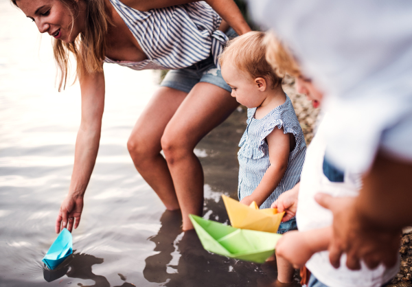 A midsection of young family with two toddler children outdoors by the river in summer, playing with paper boats.