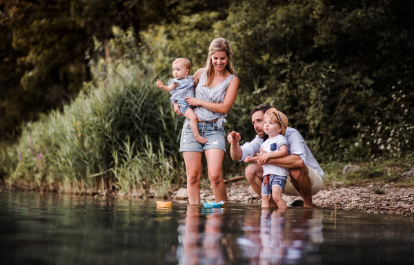 A young family with two toddler children outdoors by the river in summer, playing with paper boats.