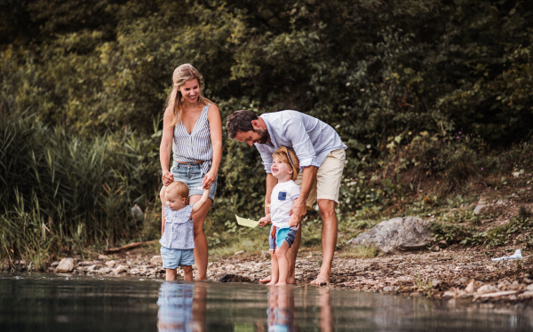 A young family with two toddler children outdoors by the river in summer, playing with paper boats.