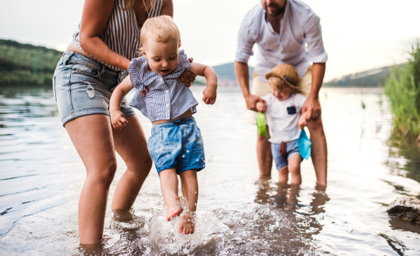 A midsection of young family with two toddler children outdoors by the river in summer, playing with paper boats.