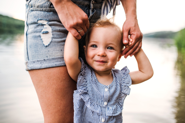 A midsection of mother with a toddler daughter standing outdoors by the river in summer.