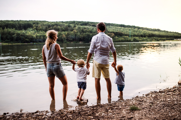 A rear view of young family with two toddler children spending time outdoors by the river in summer.