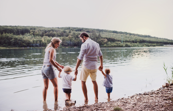 A rear view of young family with two toddler children spending time outdoors by the river in summer.