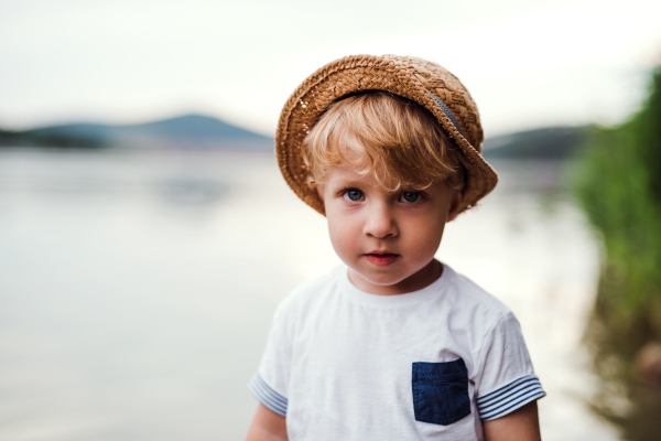 A small toddler boy standing barefoot outdoors by a river in summer, looking at camera.