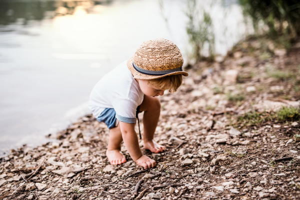 A small toddler boy standing barefoot outdoors by a river in summer, playing.