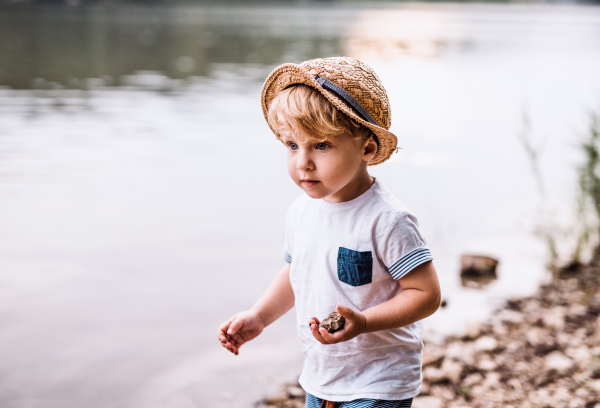 A small toddler boy standing barefoot outdoors by a river in summer, playing with rocks.