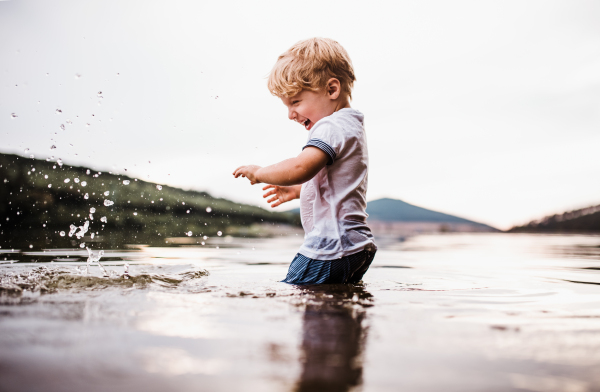 A wet, small toddler boy standing barefoot outdoors in a river in summer, playing with rocks.