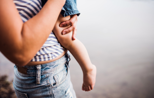 A midsection of mother with a toddler daughter standing outdoors by the river in summer.