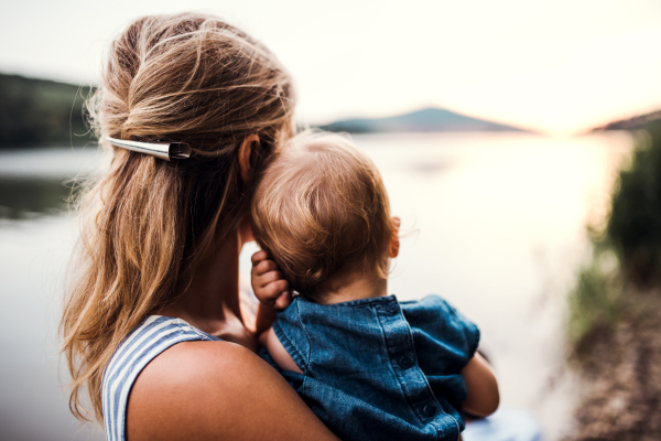 A rear view of mother with a toddler daughter standing outdoors by the river in summer.