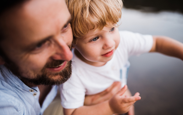 A close-up of mature father with a toddler boy outdoors by the river in summer.