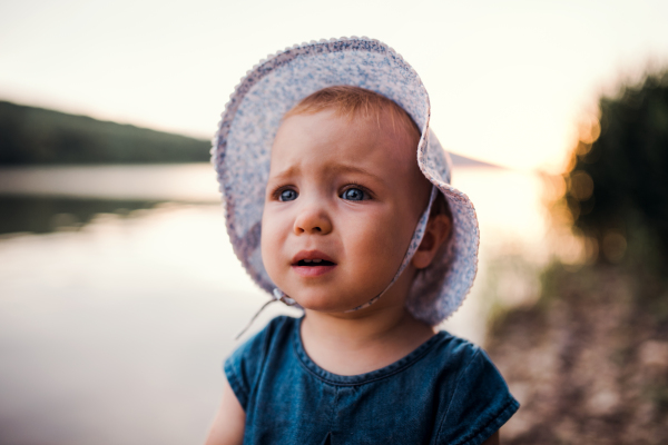 A portrait of sad small toddler girl with a hat standing outdoors by a river in summer.