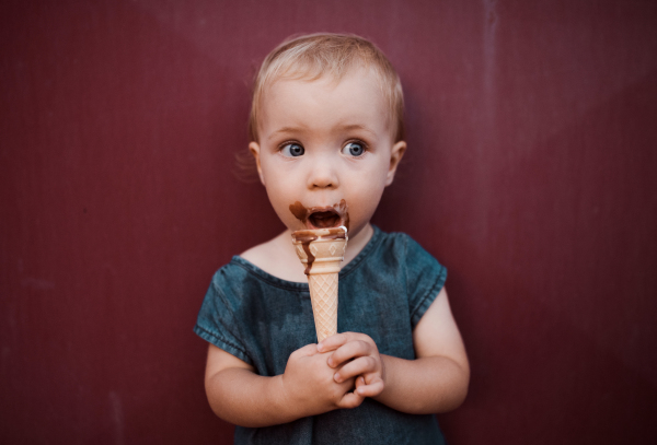 A front view of cute small toddler girl outdoors in summer, eating ice cream.