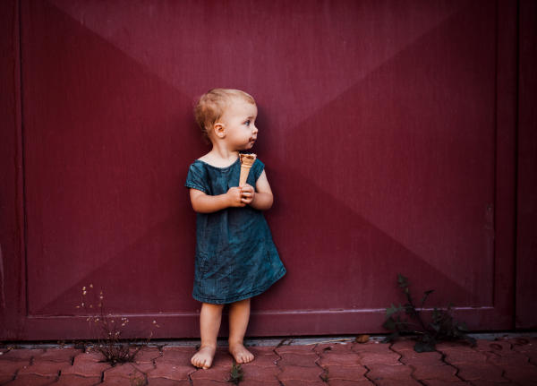 A front view of cute small toddler girl outdoors in summer, eating ice cream.